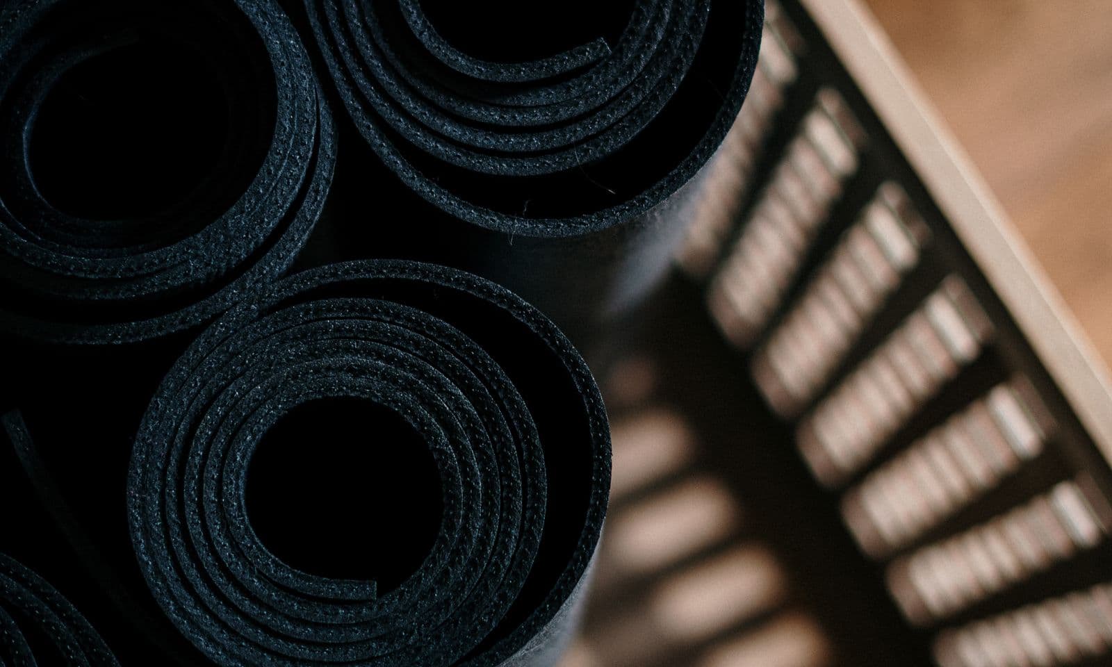 Black rolled yoga mats stacked in a woven wooden crate, top-down editorial shot with dramatic dappled shadows — the best yoga mats for teachers in 2026